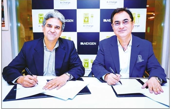 Two men in suits sit at a table and sign documents, with a NexGen Auto & ACharge logo in the background.