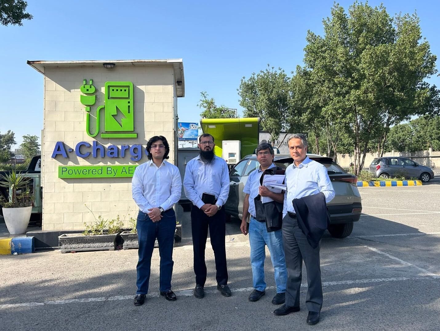 Four men stand in front of an electric vehicle charging station labeled "A-Charge" in a parking lot on a sunny day.